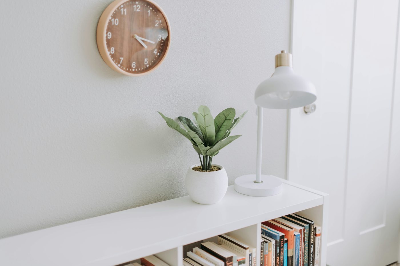 A calm, minimalist white workspace: a leafy green plant in a white pot and a white study lamp on a white shelf, with a wooden wall clock above and books on a lower shelf.