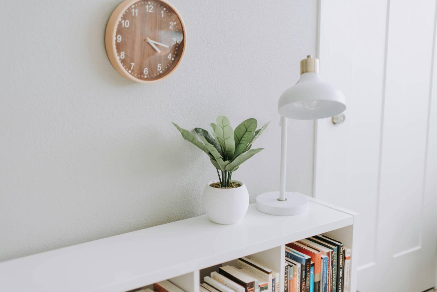 Minimalist bookshelf, white wall, brown timber clock with white markings on the wall. A white bookshelf with some books on the shelves and a white lamp on top, next to a white pot plan with a green leafy plant in it.